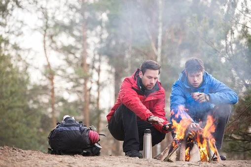 Two men in outdoor jackets sitting by a campfire in a forest, with a backpack and water bottle nearby.