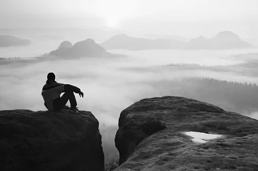 A person sitting on a rock ledge overlooking a foggy mountain landscape in black and white.