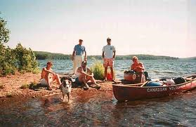 A group of five people by a lake, with three sitting on the shore and two standing nearby, and a boat on the water.