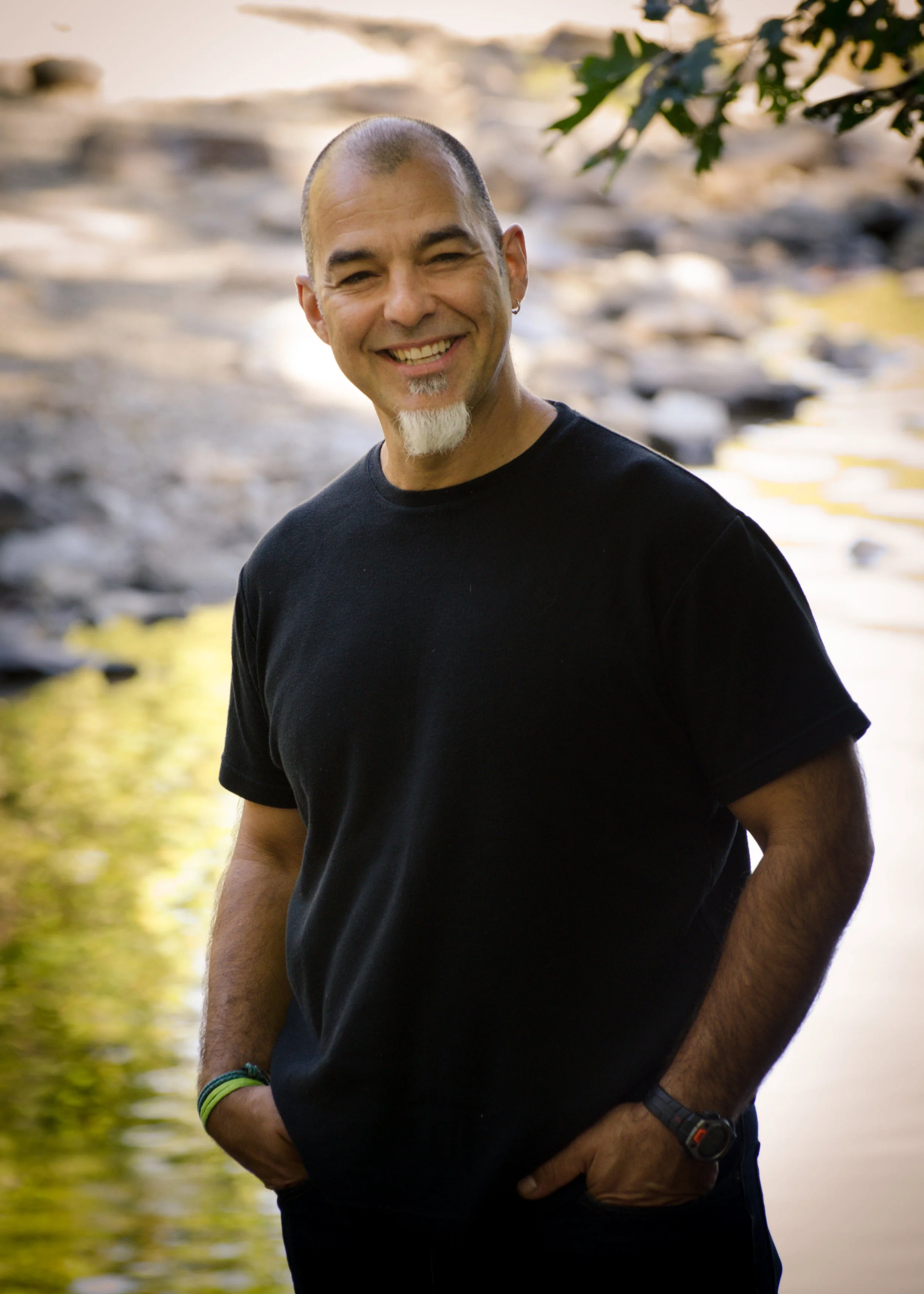 A smiling man with a goatee and earring, standing outdoors near a river, wearing a black t-shirt, with trees and rocks in the background.