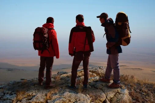 Three hikers with backpacks standing on a rocky hilltop overlooking a flat landscape.