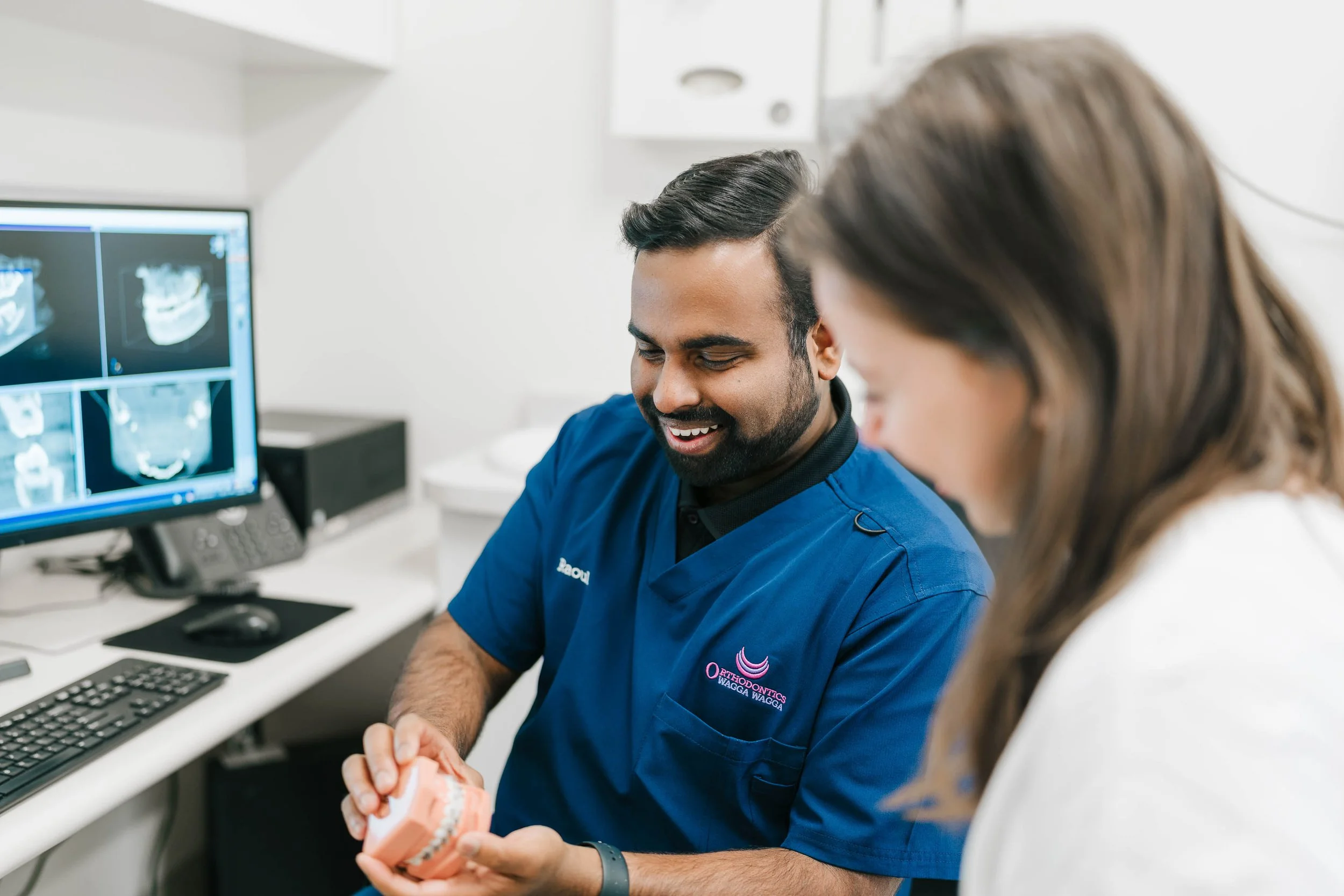 A dental professional wearing a blue scrubs holding a dental model while talking to a patient in a clinic with computer screens displaying dental x-rays.