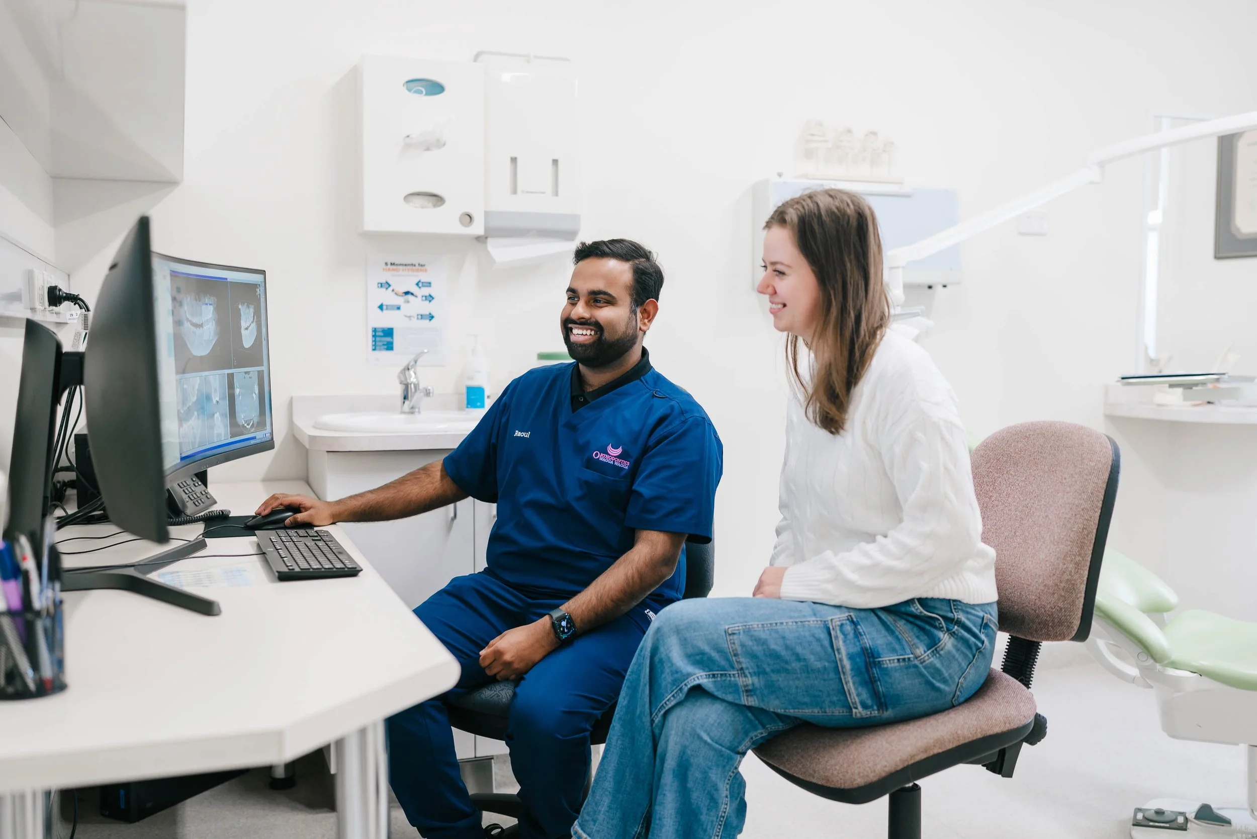 A dentist and a female patient sitting together in a dental clinic, looking at computer screens displaying dental X-rays, both smiling during a consultation.