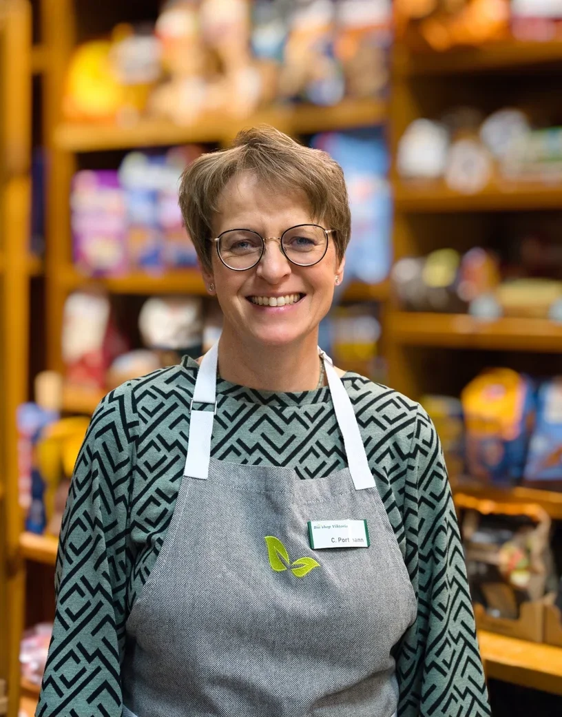 Lächelnde Frau mit Brille und grauem Apron in einem Supermarkt, umgeben von Regalen mit Lebensmitteln.