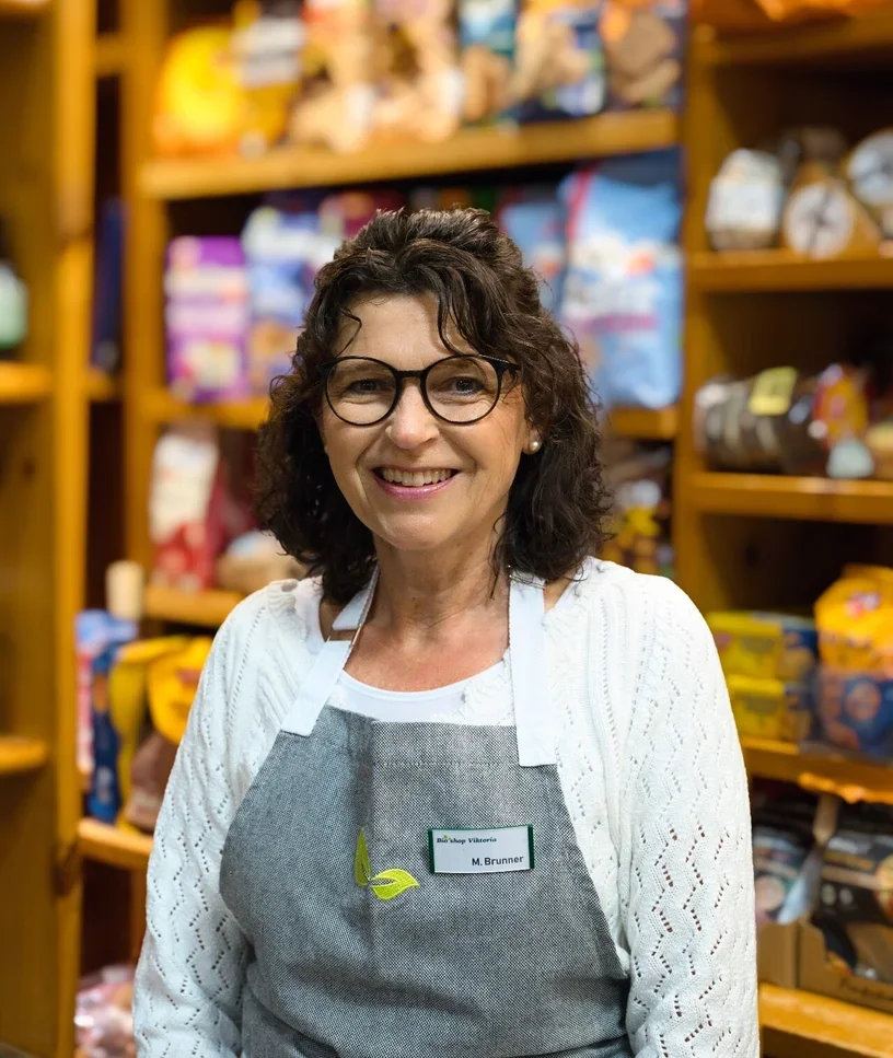 Eine Frau mit Brille und lockigem Haar, die in einem Laden steht, umgeben von Regalen mit bunten Produkten, trägt eine graue Schürze und lächelt.