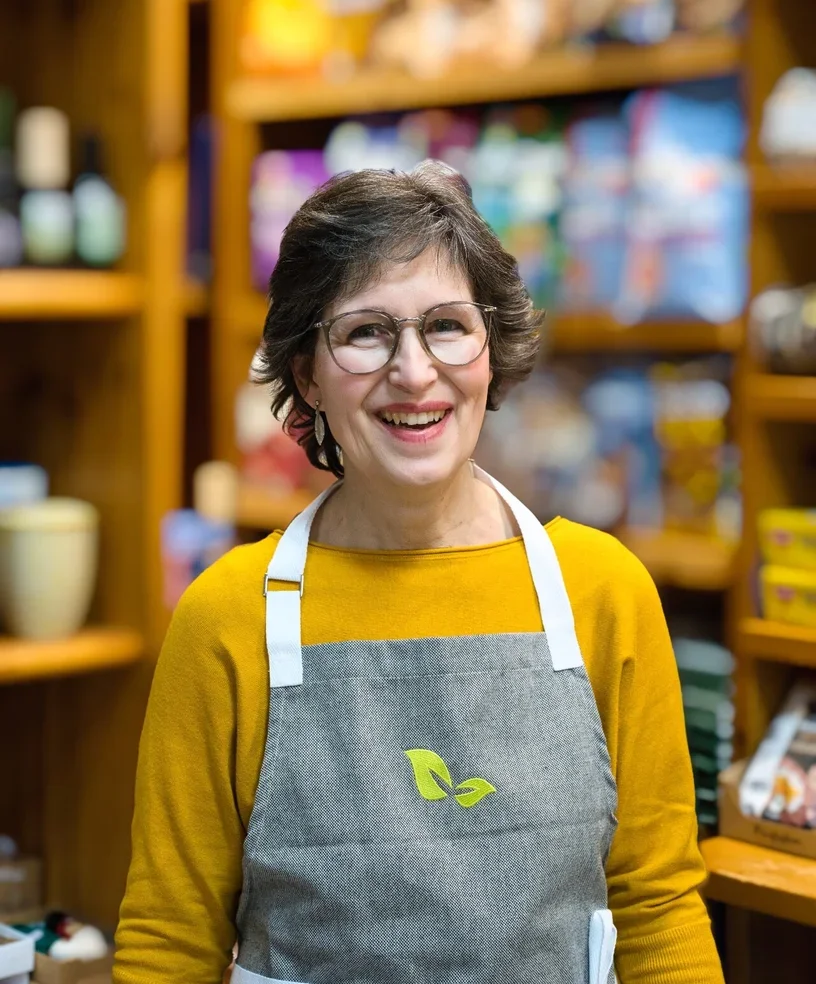 Eine smiling Frau in einem Laden, trägt eine graue Schürze mit einem grünen Blattsymbol, umgeben von Garten- und Haushaltsprodukten auf Holzwänden.