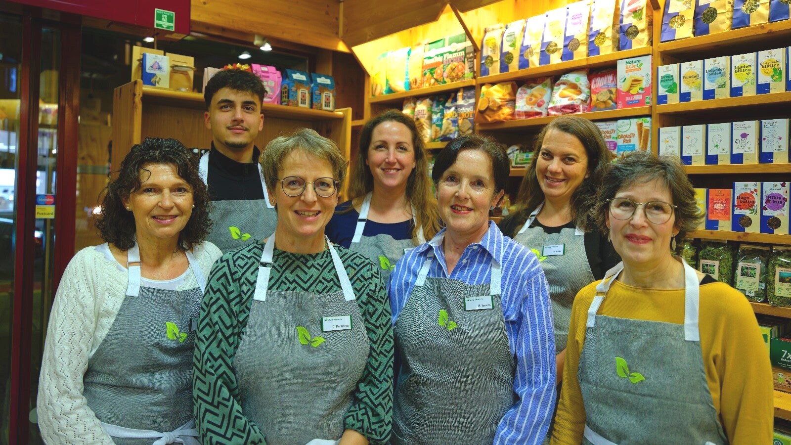Eine Gruppe von acht Frauen und einem Mann steht in einem Laden mit Holzregalen voller bunter Verpackungen und Produkten, alle tragen graue Schürzen mit einem grünen Logo von Blättern.