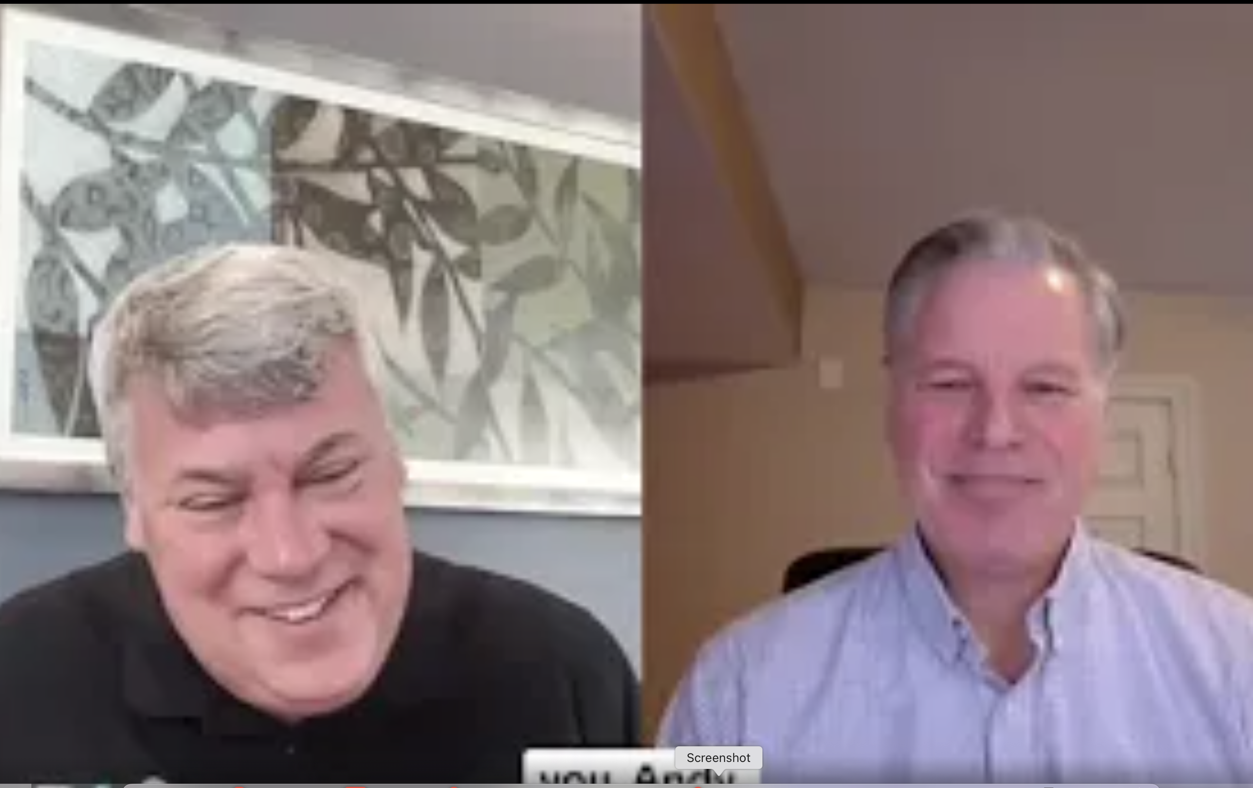 Two middle-aged men are engaged in a video call, both smiling. The man on the left has light-colored hair and is wearing a black shirt, with a patterned art piece in the background. The man on the right has short gray hair and is wearing a light-colored button-up shirt, with a solid-colored wall and a door in the background.