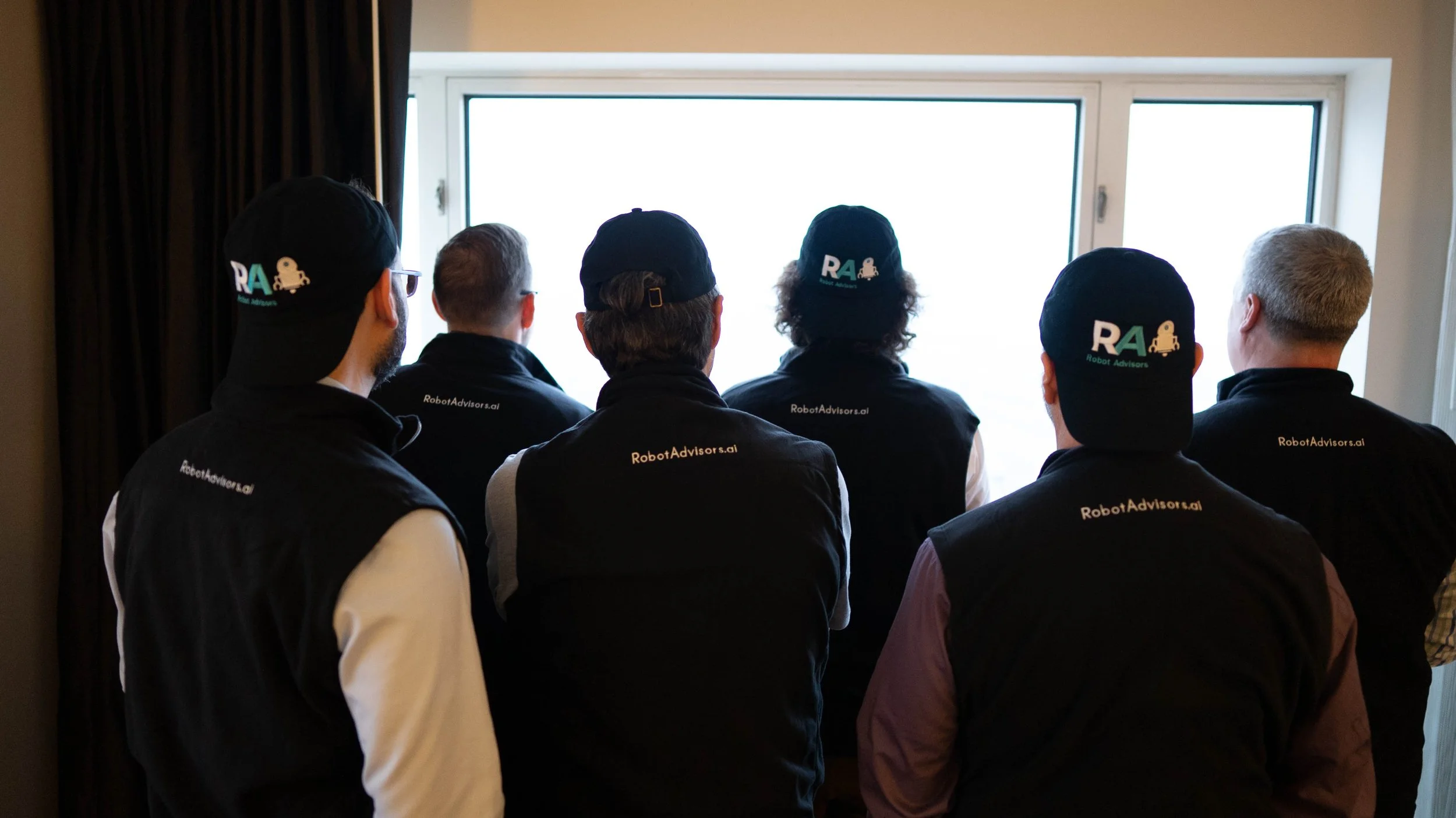 Six individuals wearing black jackets and hats with the 'Robot Advisors' logo, sitting and watching a bright window