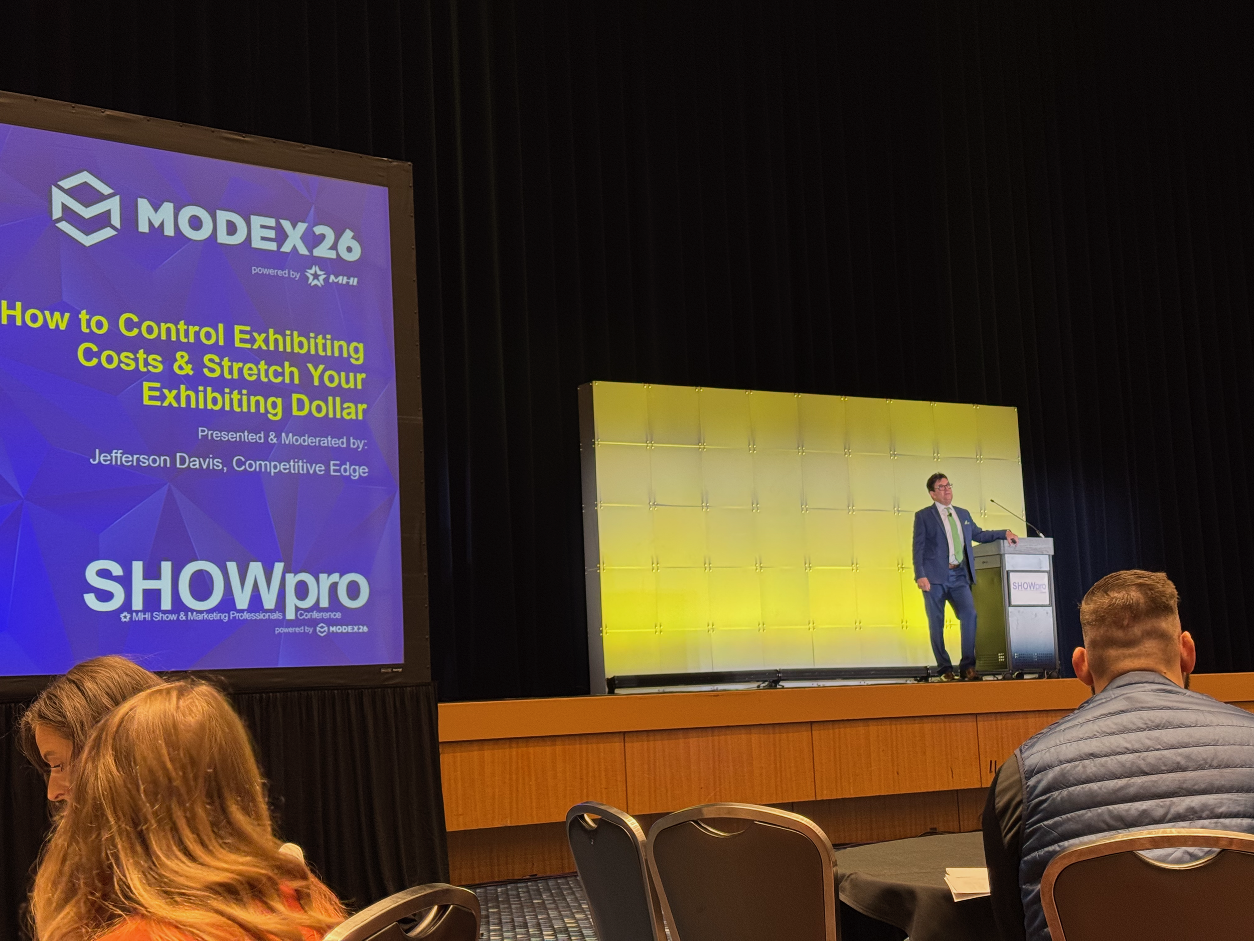 A man in a blue suit stands at a podium on a stage, giving a presentation at the MODex26 conference. The presentation slide reads 'How to Control Exhibiting Costs & Stretch Your Exhibiting Dollar' and features the SHOWpro and MODex26 logos. The audience members are seated facing the stage.