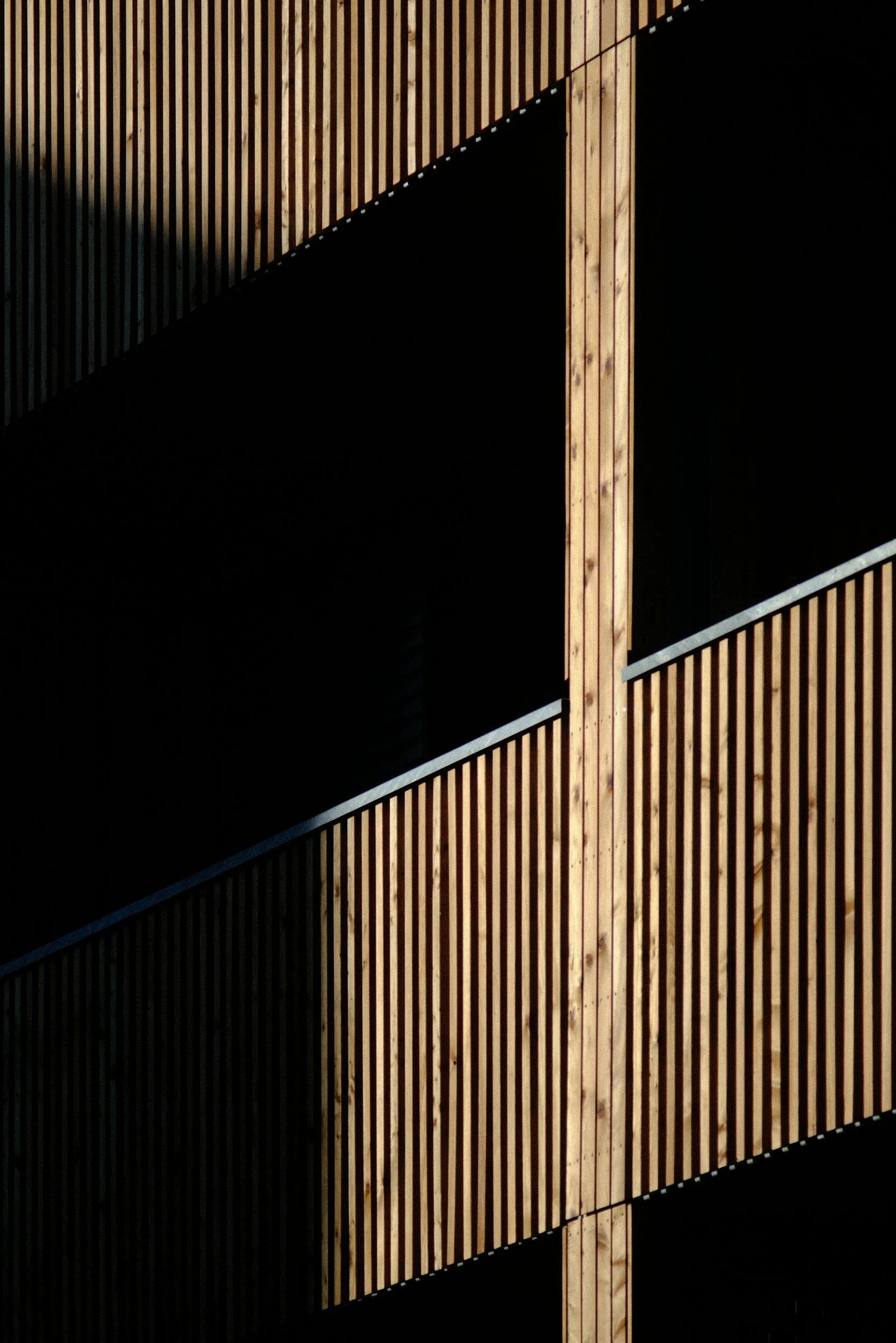 Close-up of a modern building with wooden vertical slats on the exterior, creating a geometric pattern with contrasting shadowed and illuminated sections.