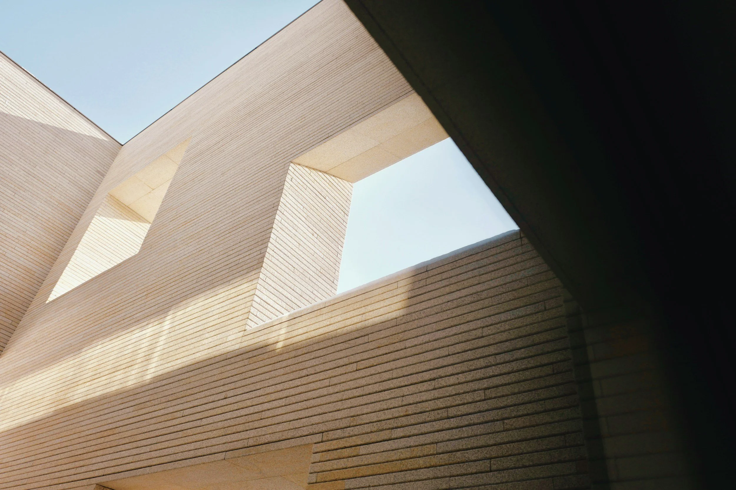 Low-angle view of modern building with beige brick walls and large rectangular openings, under a clear blue sky.