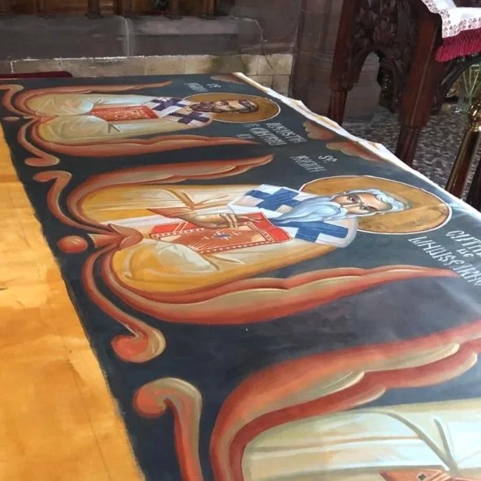 A large religious banner or poster laid out on a table, depicting saint-like figures with halos, colorful robes, and holding religious items, surrounded by ornate decorative elements.