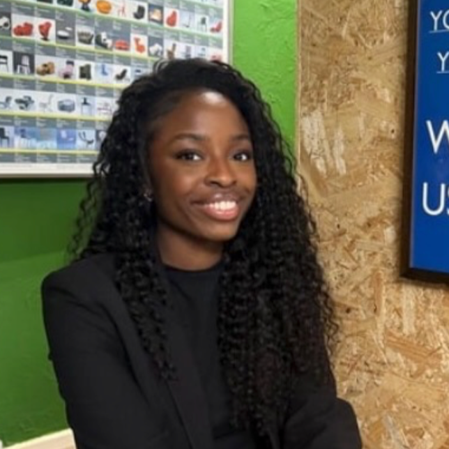 A young woman with dark, curly hair smiling at the camera, wearing a black blazer, standing indoors with a green wall and a bulletin board in the background.