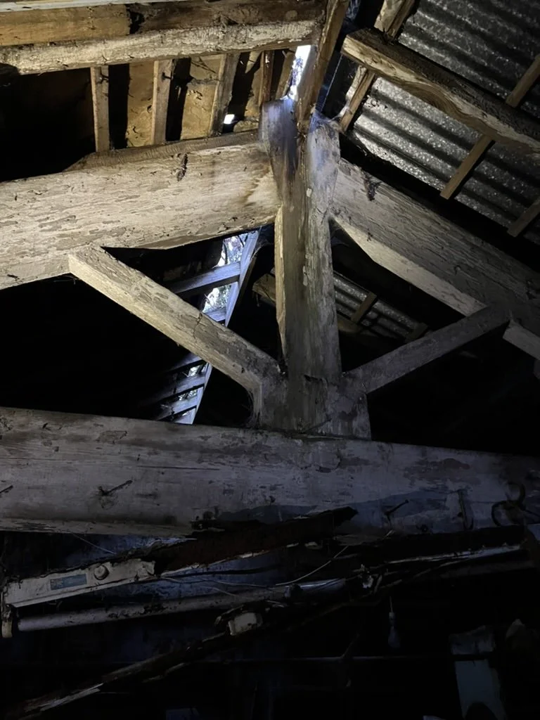 Close-up of the attic structure showing aged, weathered wooden beams and rafters, with some sections covered in corrugated metal roofing. Natural light filters through gaps in the roof.