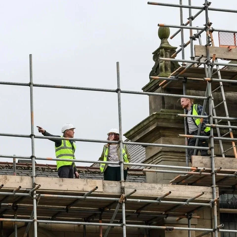 Three construction workers, two men and one woman, wearing safety vests and hard hats, are standing on scaffolding on a roof, discussing and inspecting the work, with a chimney in the background.