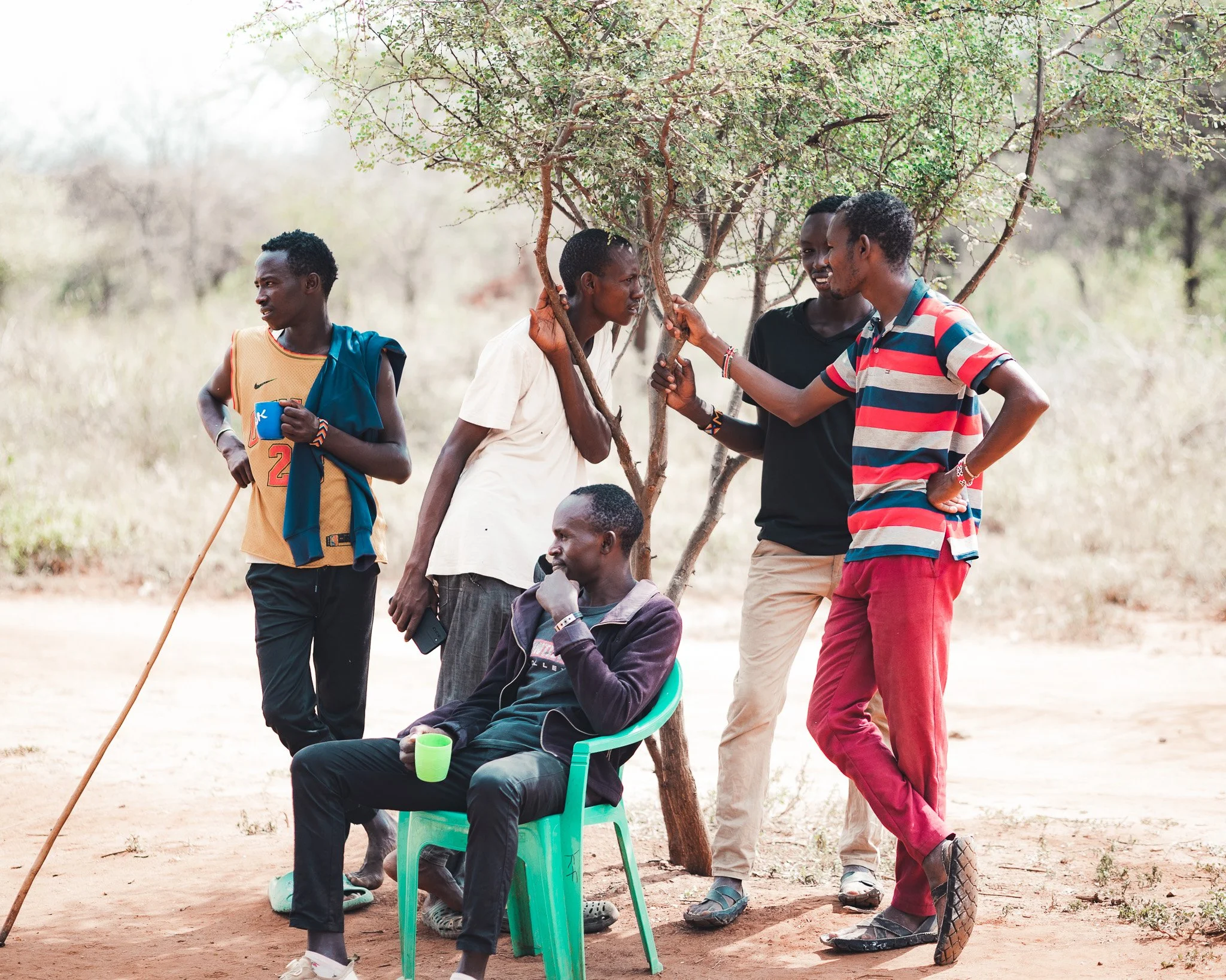 Four young people are gathered outdoors under a small tree on Kijani Farm. One woman is sitting on a bright green chair, holding a green mug, while three others stand nearby and converse. The setting is a dry, open area with sparse greenery.