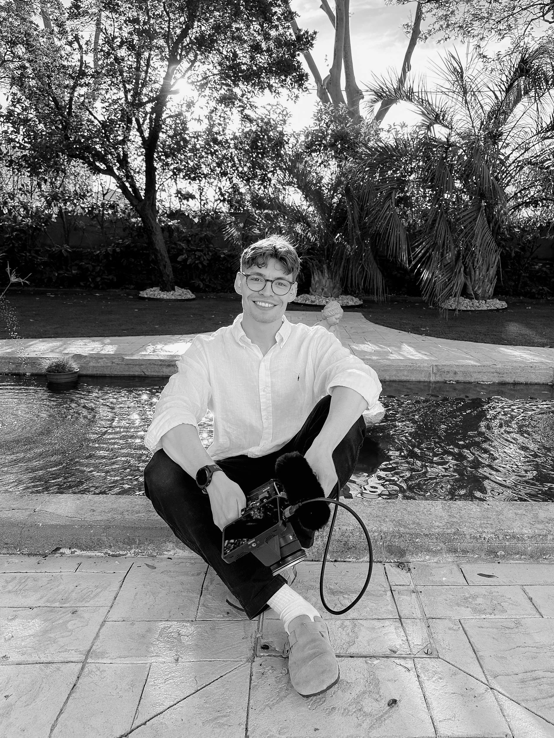A young man sitting by a pool with a camera hanging around his neck, smiling at the camera. The photo is taken outdoors with trees and plants in the background, black and white.