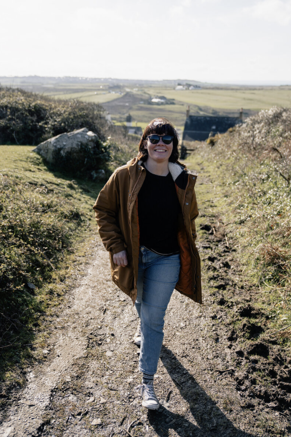 Woman wearing sunglasses, a brown jacket, black shirt, and jeans walking on a dirt path in a rural landscape with green hills and farm buildings in the background.