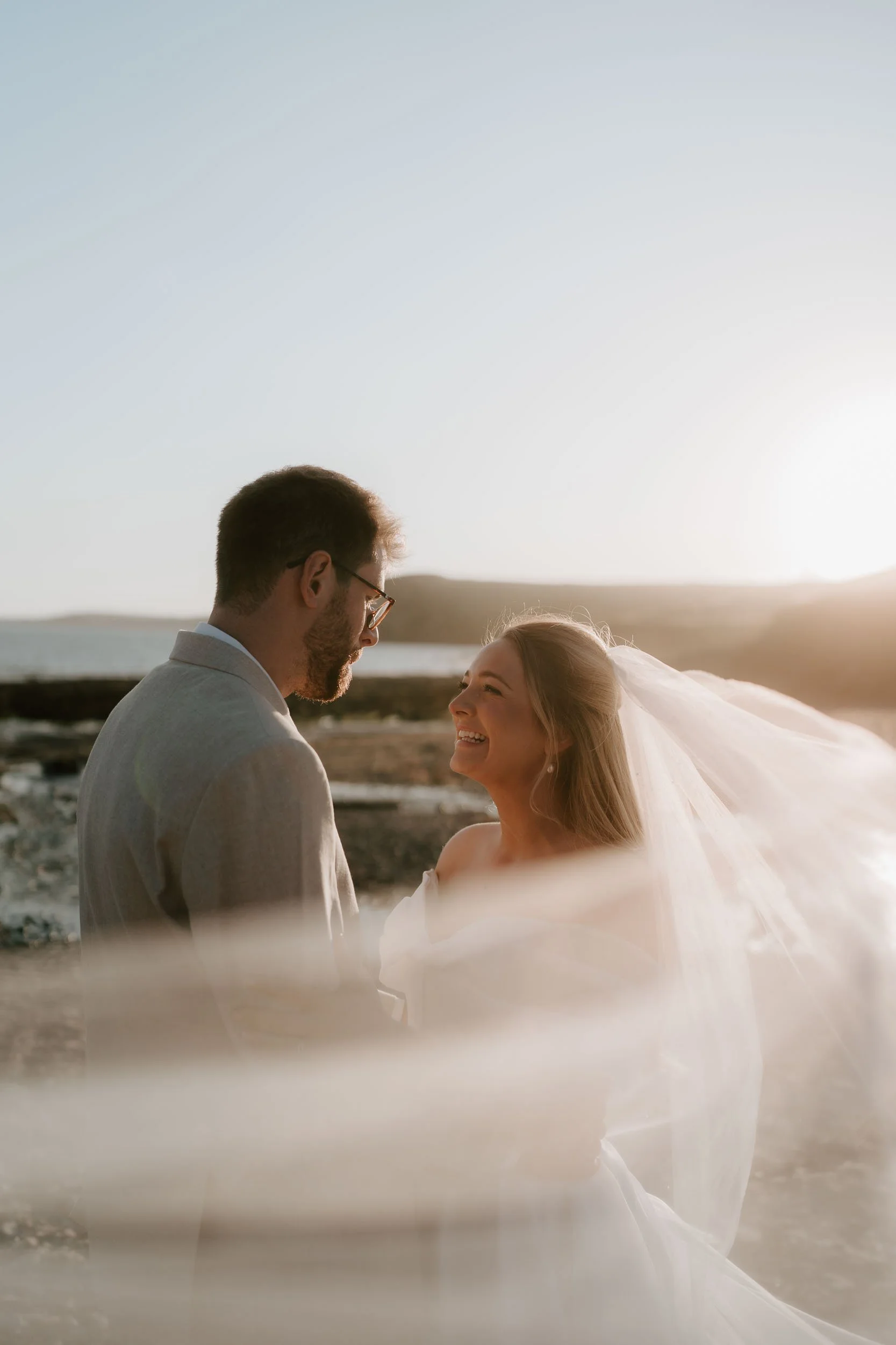 A couple in wedding attire, standing close together outdoors at sunset, smiling and looking into each other's eyes, with the ocean and sky in the background.