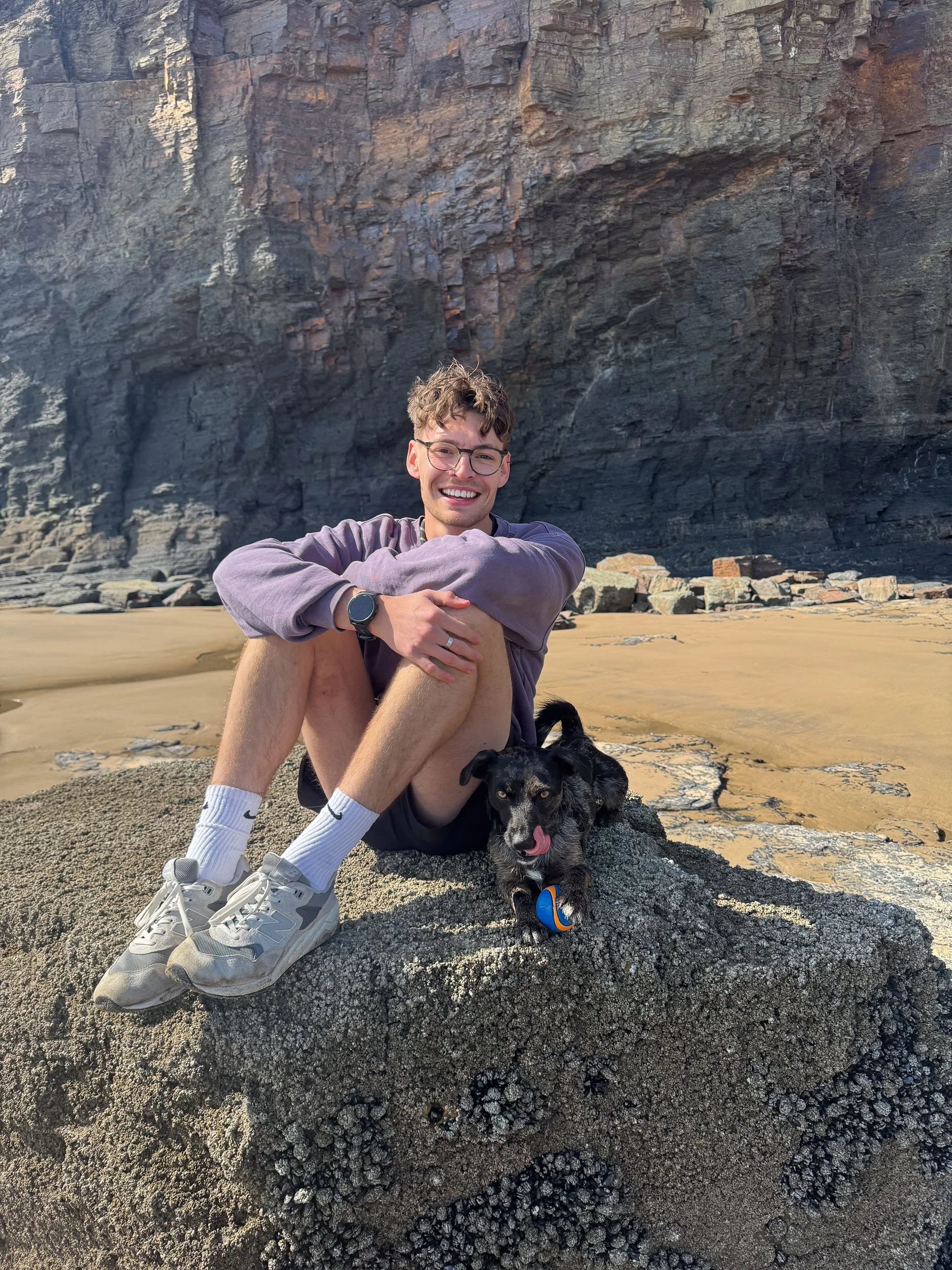 A young man with glasses, curly hair, and a wide smile sits cross-legged on rocks at the beach, hugging his knees, with a black and white dog lying beside him. Behind them, a large rocky cliff towers over the sandy shore.