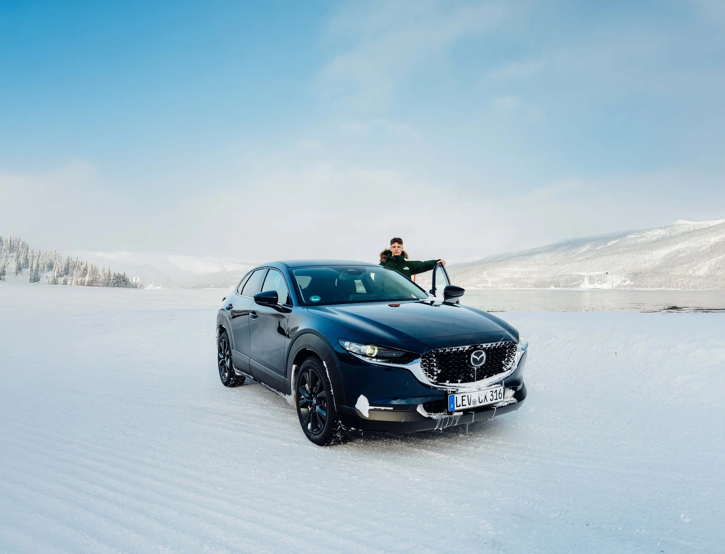 A young man standing beside a black Mazda SUV on snow-covered ground in a winter landscape with snow-covered trees and mountains.