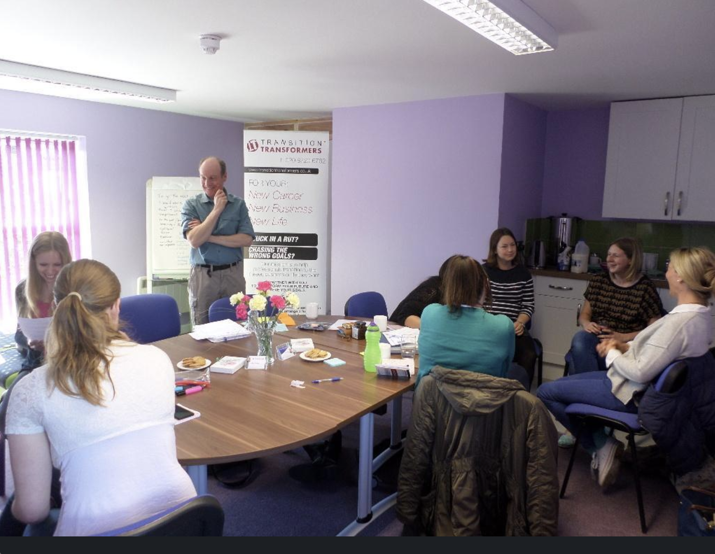 Ross standing by a white board with a table and several women discussing topics