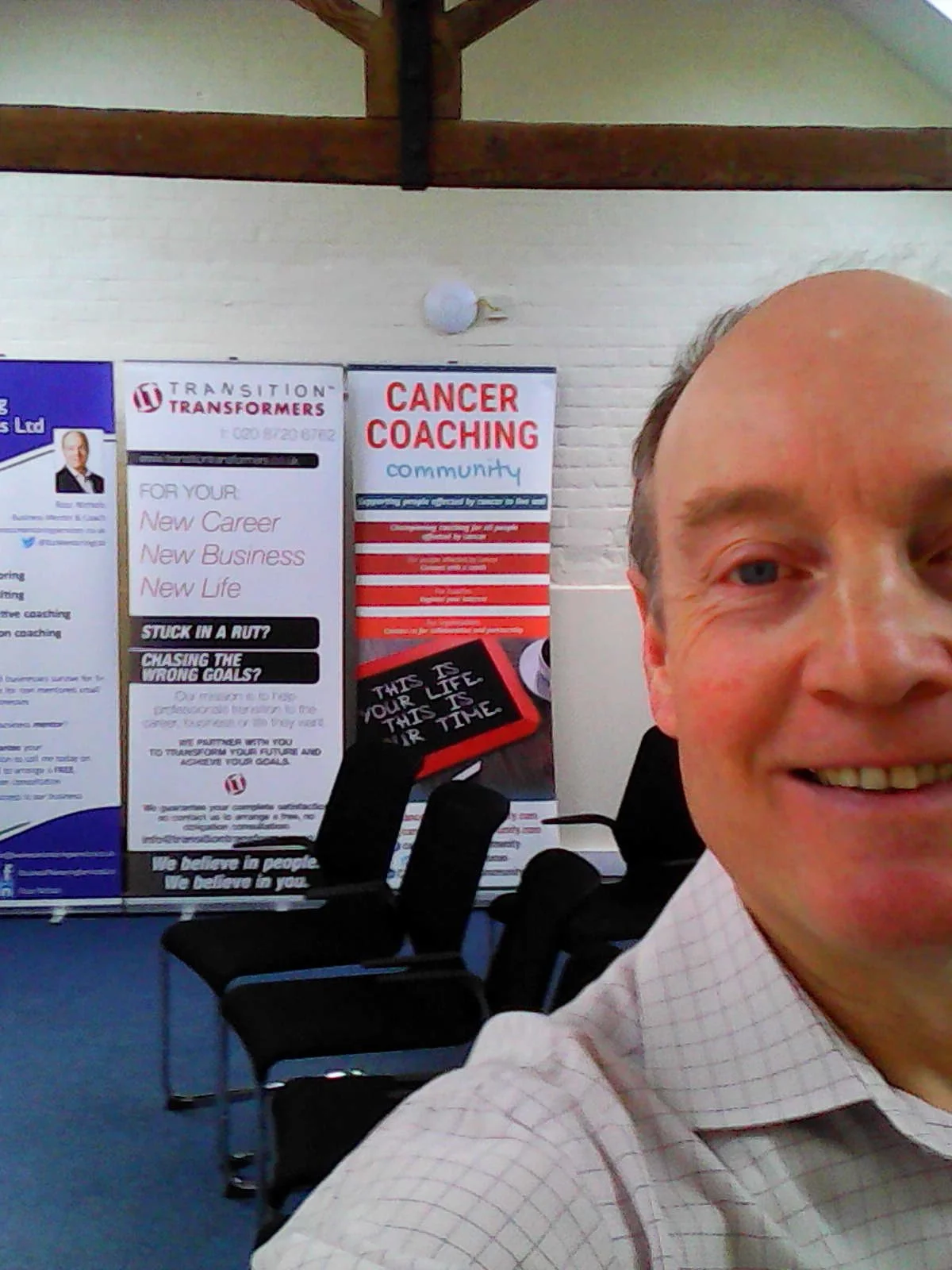 Ross Nichols, in a checkered shirt, smiling at a camera, with posters behind him promoting career coaching and community cancer coaching as part of Salisbury International Coaching Week.