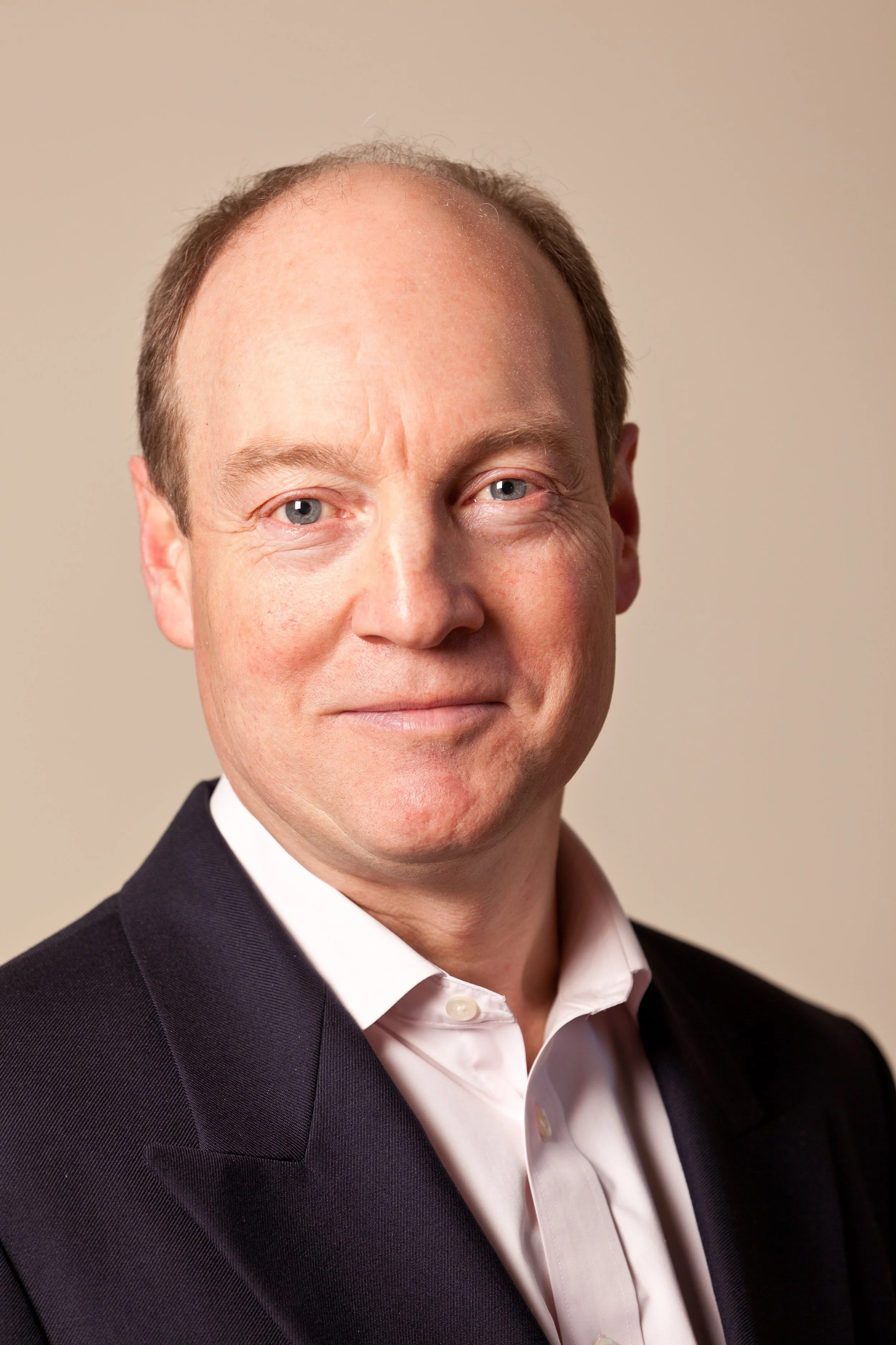 A close-up portrait of Ross Nichols with fair skin, blue eyes, and short light brown hair. He is wearing a dark suit jacket and a white dress shirt, smiling subtly against a neutral background.