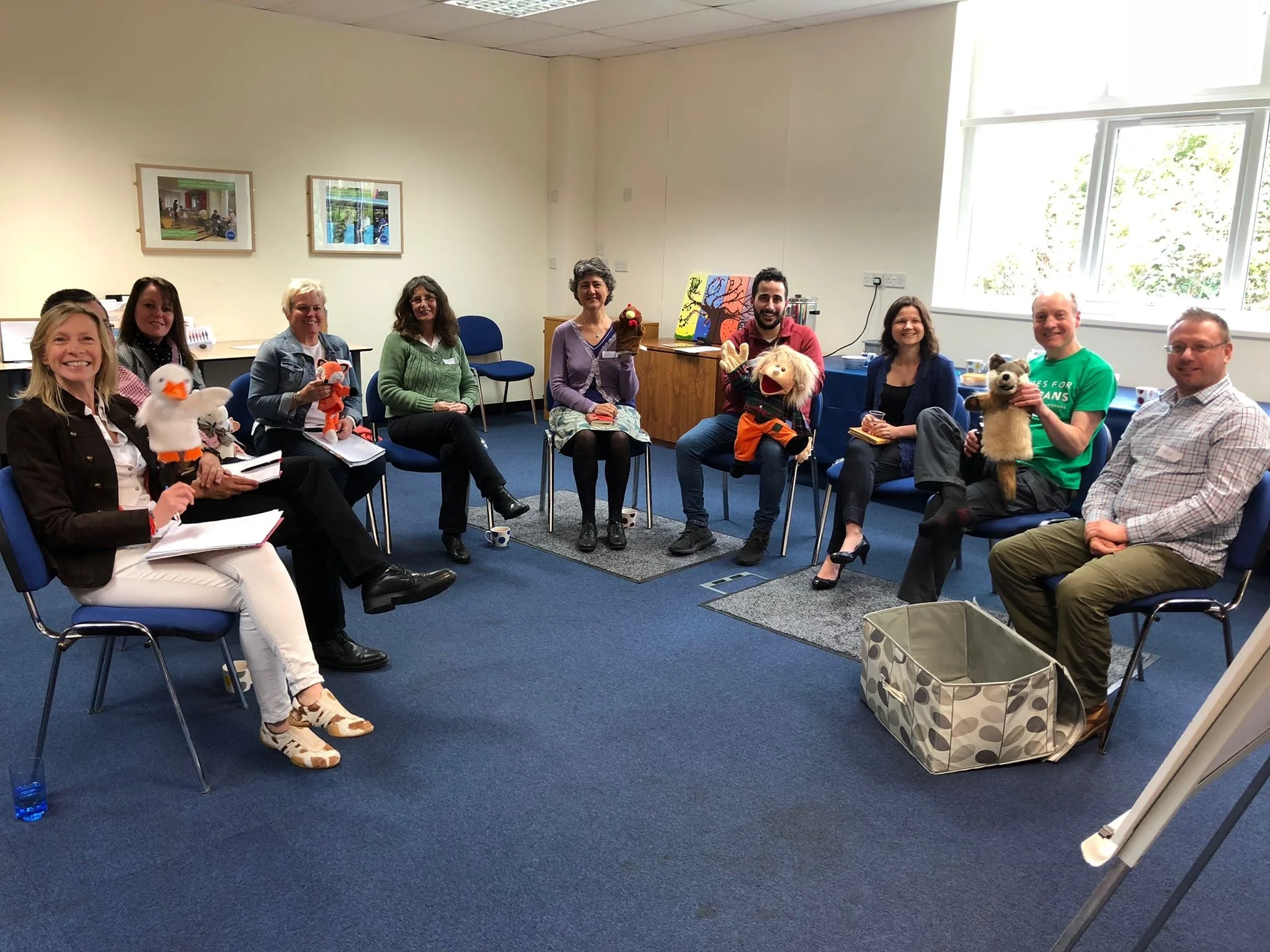 A group of nine people, including Ross Nichols, sitting in a semi-circle in a room, some holding puppets as part of the organising of the Salisbury International Coaching Week