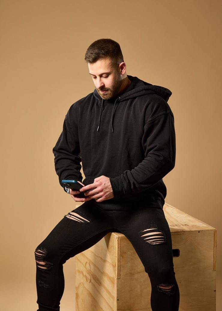 A man with short dark hair and a beard, wearing a black hoodie and ripped black jeans, sitting on a wooden box and looking at his phone against a plain tan background.