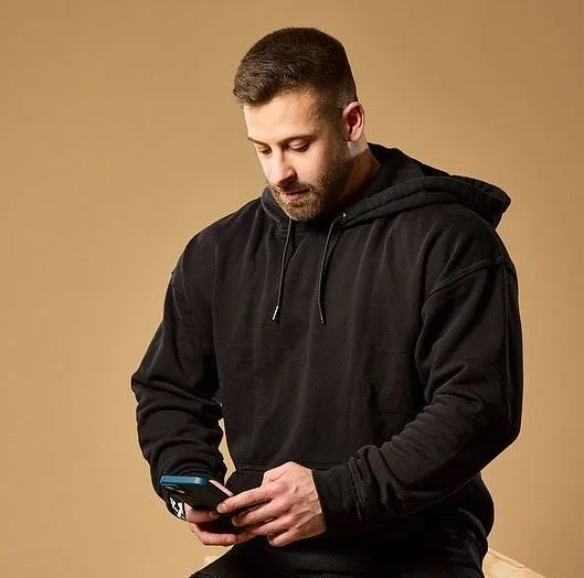 A man with short dark hair and a beard sitting against a tan background, wearing a black hoodie, looking down at his phone.