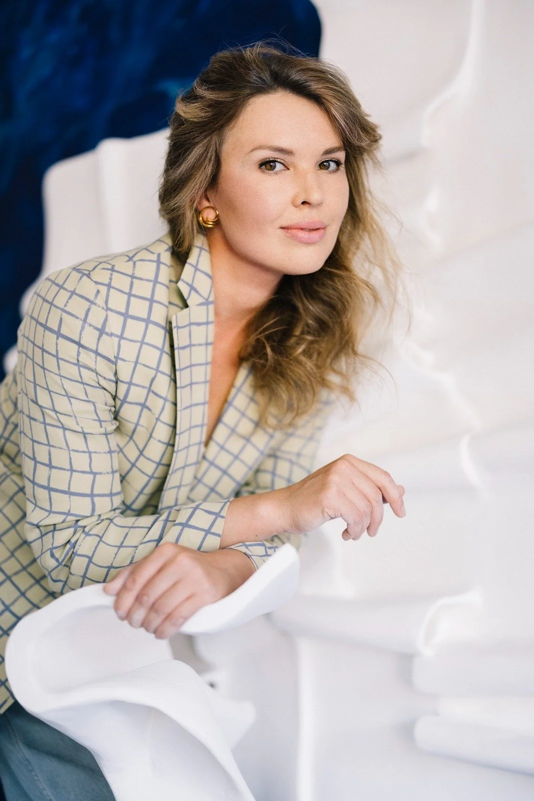 A woman with wavy hair and gold earrings sitting on a white chair with a white textured wall in the background.