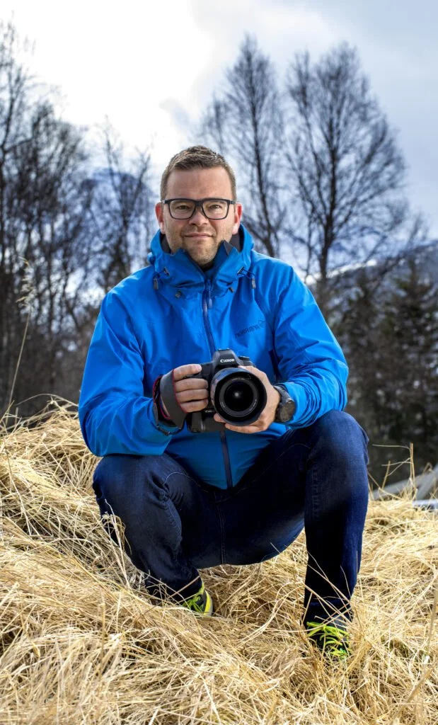 En mann i blå jakke som tar et bilde med et digitalt kamera, satt på gress omringet av trær uten løv foran en blå himmel.