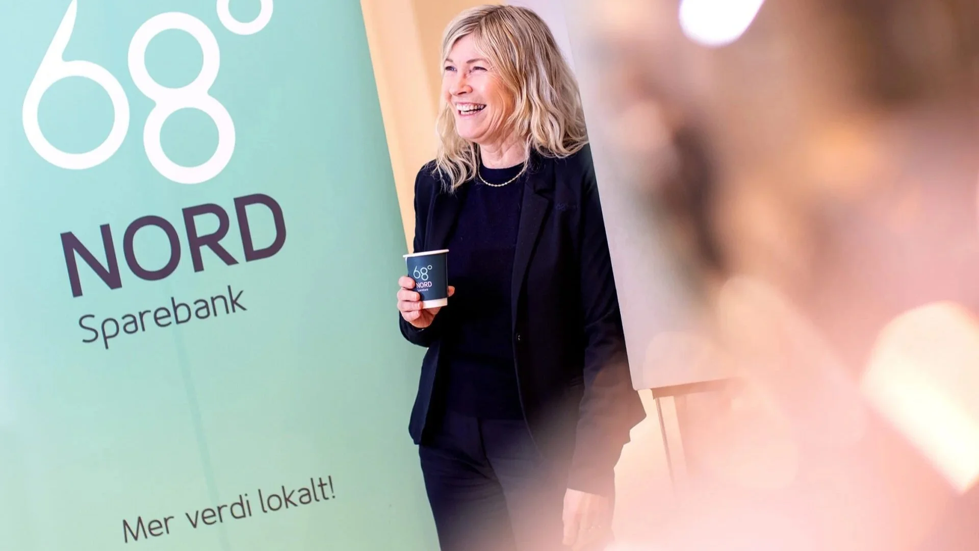En kvinne smiler og holder en kopp på en konferanse eller markering for Nord Sparebank. Bakgrunnen har teksten «68° Nord» og noen andre ord. Det er en innendørs setting med uklar bakgrunn.