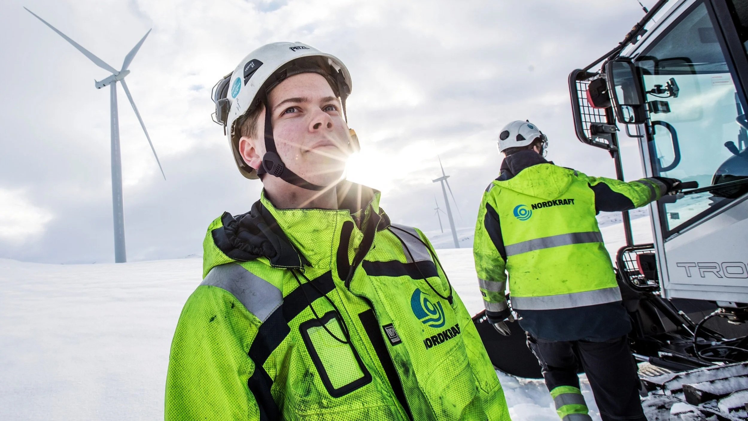 To personer i høye synlige jakker med logoet «Nordkraft» står i snødekt landskap med vindmøller i bakgrunnen, en av dem ved en maskin.»