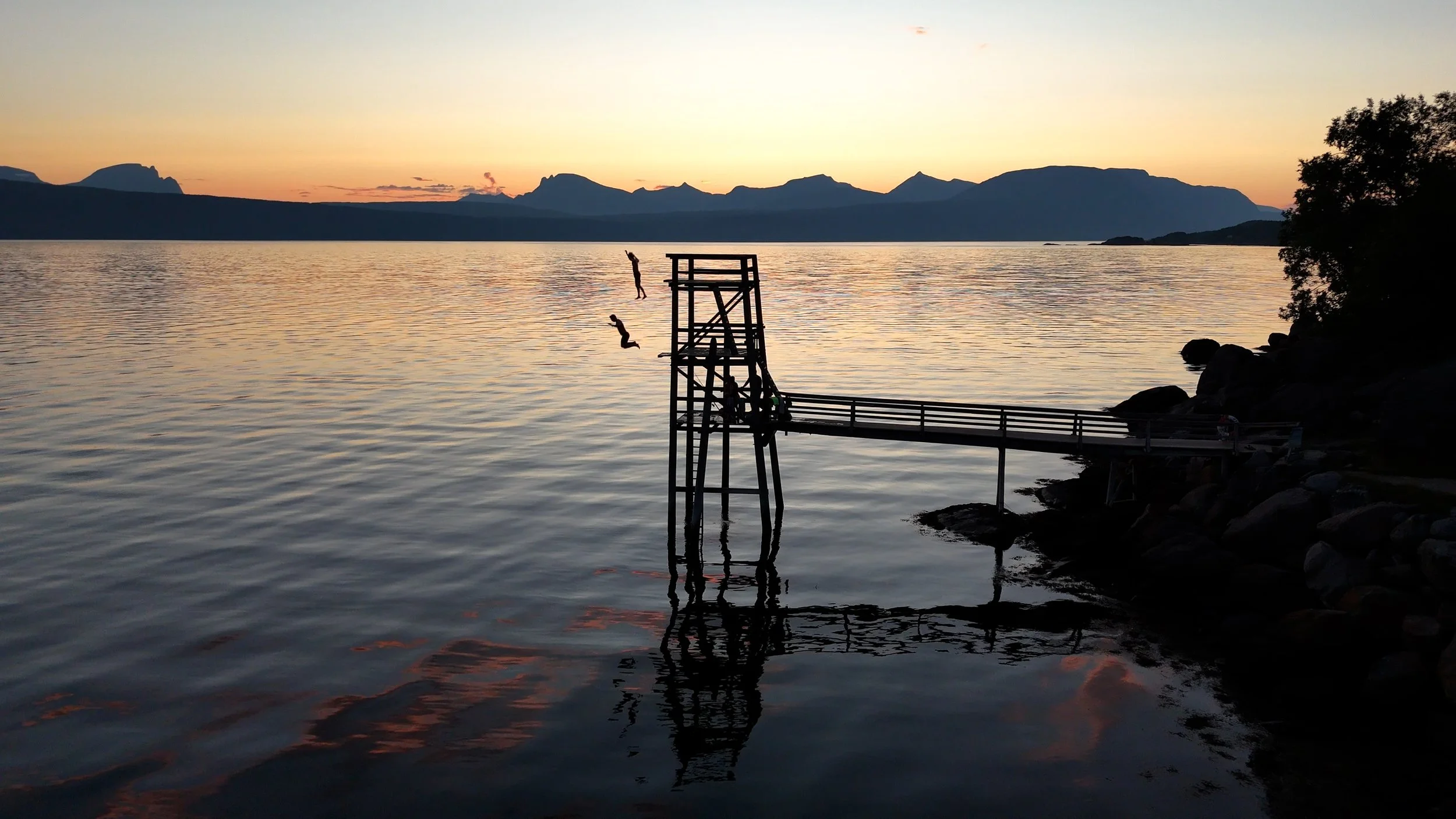 Siluetter av barn som hopper ut fra en flytende plattform på en rolig innsjø under en solnedgang med fjell i bakgrunnen.
