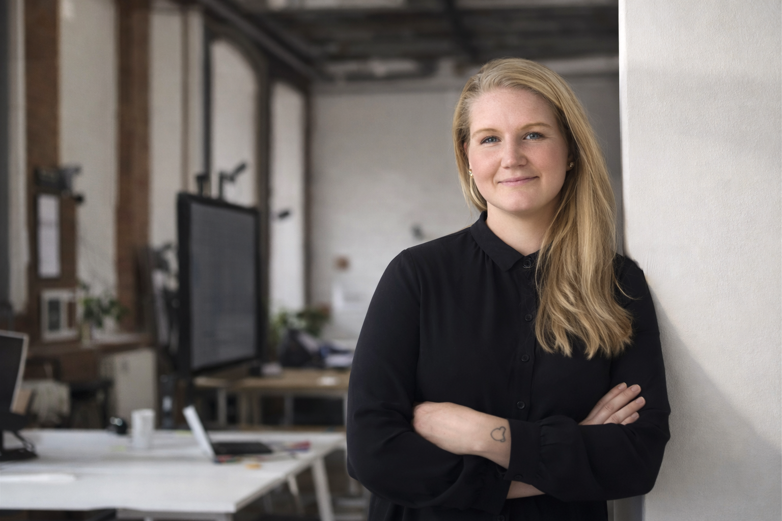 Kvindelig person står med armene over kors og smiler i et kontormiljø.