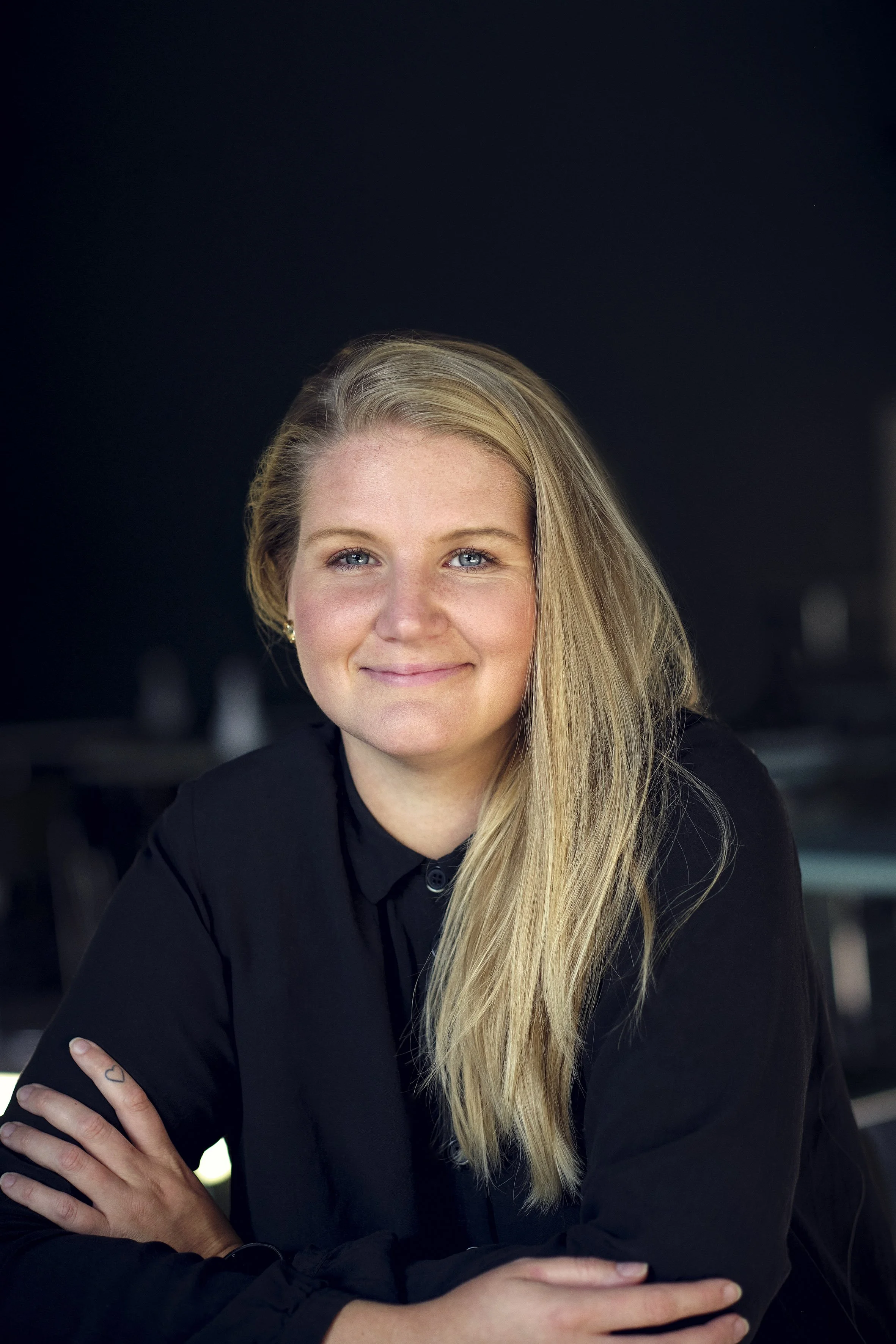 Et portræt af en smilede blond kvinde med blå øjne, iført sort tøj, siddende med armene krydsede, foran en mørk baggrund.