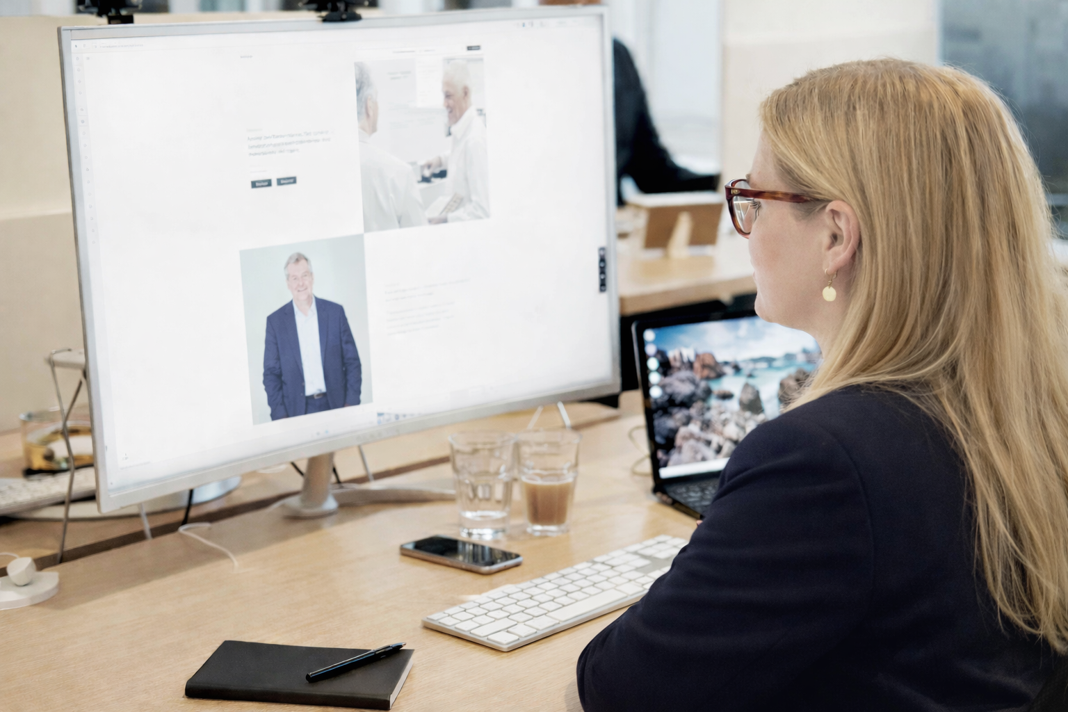 Kvinde med briller sidder foran en computerskærm, der viser billeder af folk i et kontormiljø.