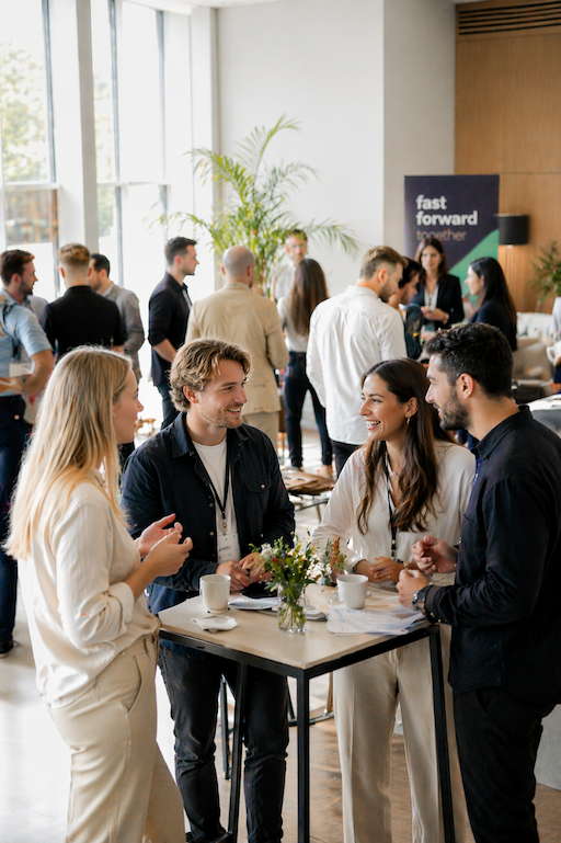 Gruppe unge voksne snakker og smiler ved en høj bord i en moderne, lyse konferencelokale med store vinduer.