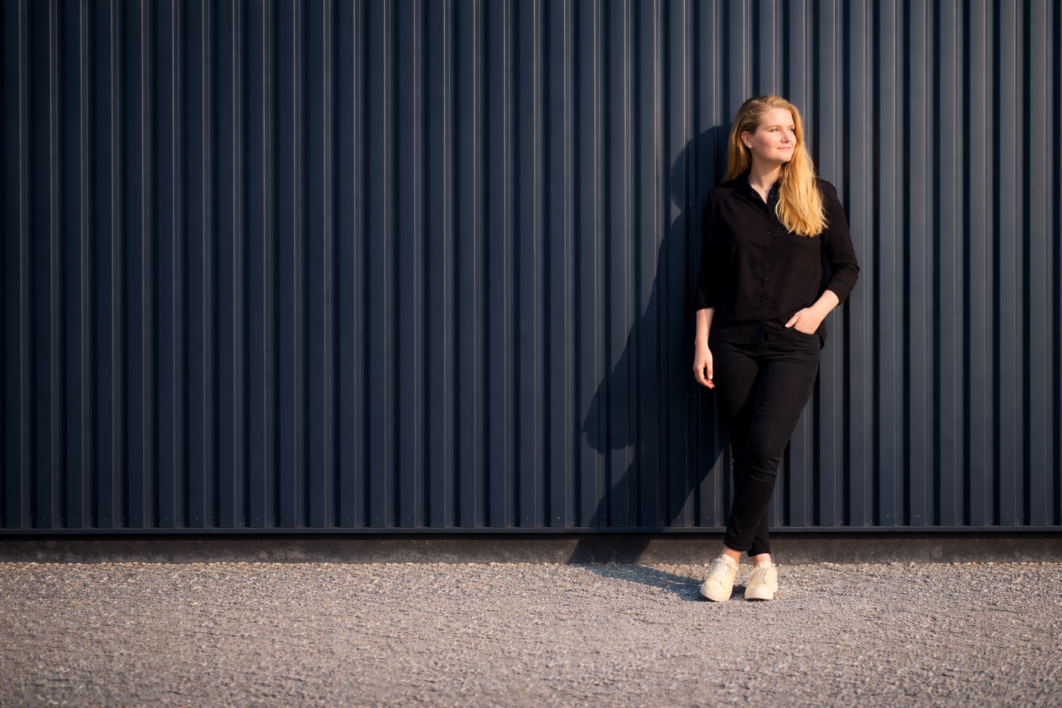 En ung kvinde i sort tøj står og holder en hånd i lommen, mens hun står foran en mørk blå væg.