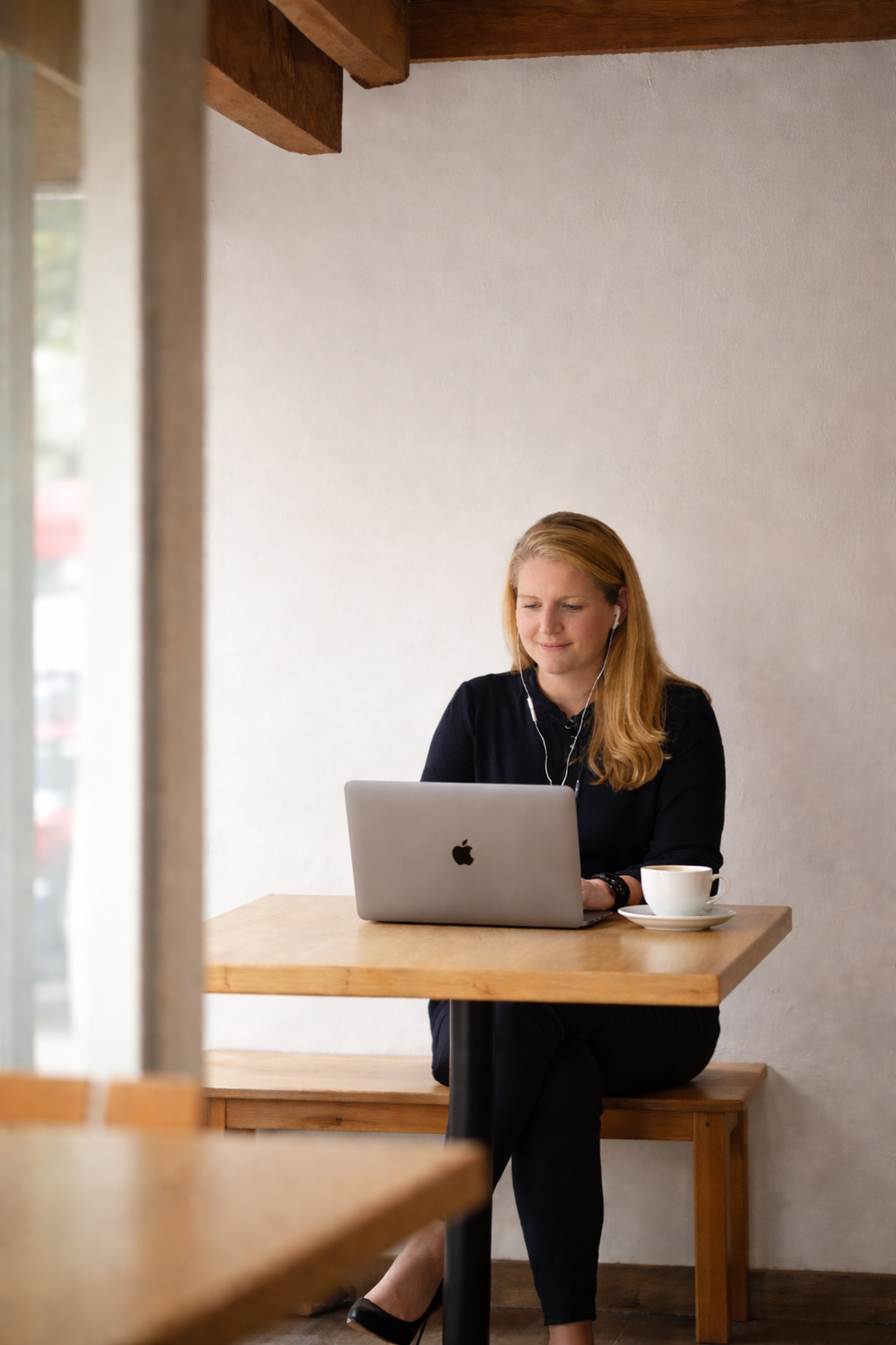 En kvinde med blondt, langt hår sidder ved et cafénbord med en MacBook, en kop kaffe og høretelefoner, smiler let og ser ned mens hun arbejder.