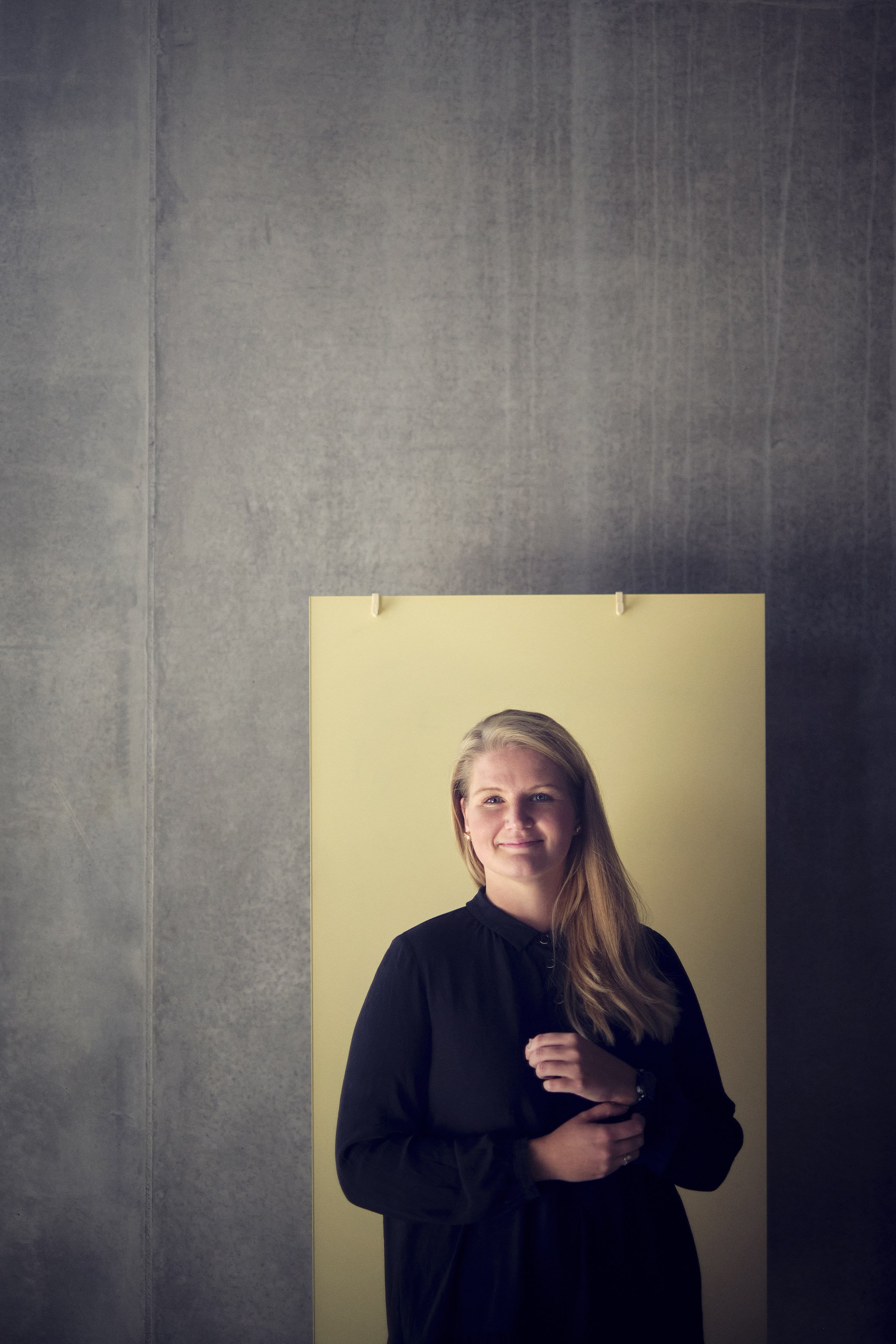 En kvinde med langt blond hår iført sort tøj, stående foran en gul baggrund, mod en grå væg, smilende mod kameraet.
