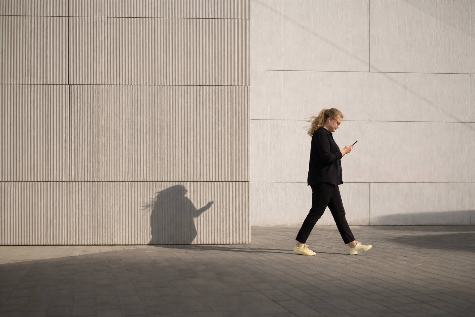 En kvinde i sort tøj går langs en hvid væg og bruger sin mobiltelefon.