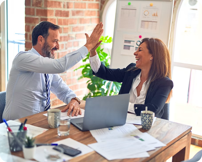 Two professionals, a man and a woman, smiling and giving each other a high five in an office setting, with documents, a laptop, and a whiteboard in the background.