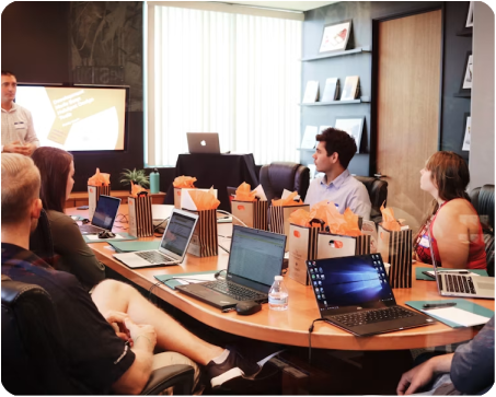 Business meeting in a conference room with people seated around a table using laptops, and a presenter standing near a large screen.
