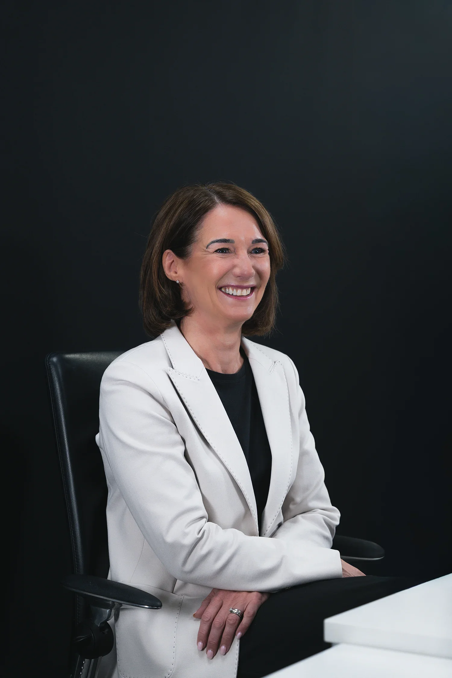 A woman sitting in a black office chair, smiling, with a white jacket and black top, against a black background.
