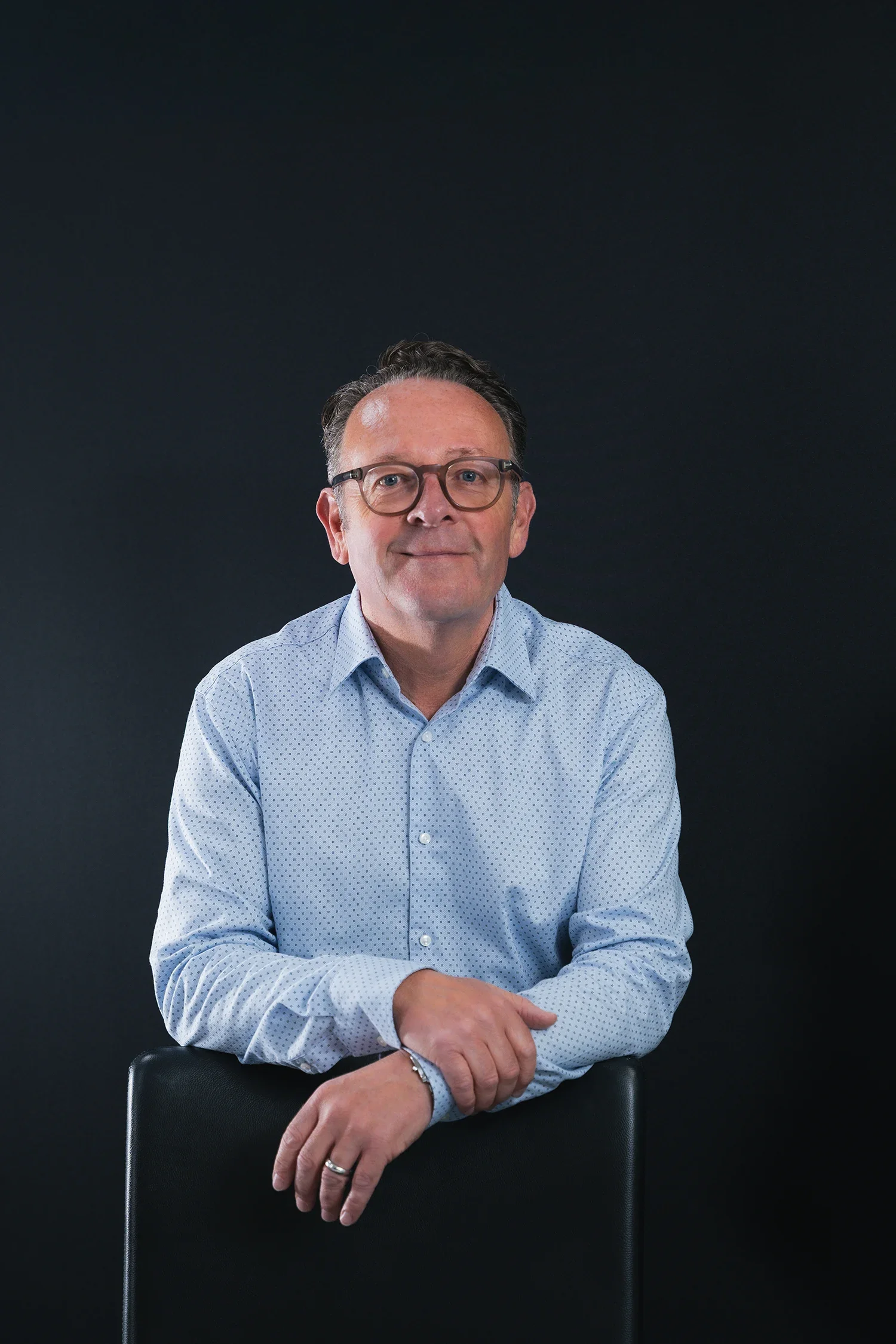 A middle-aged man with glasses is sitting and leaning on the backrest of a chair, wearing a light blue collared shirt with small polka dots, against a dark, plain background.