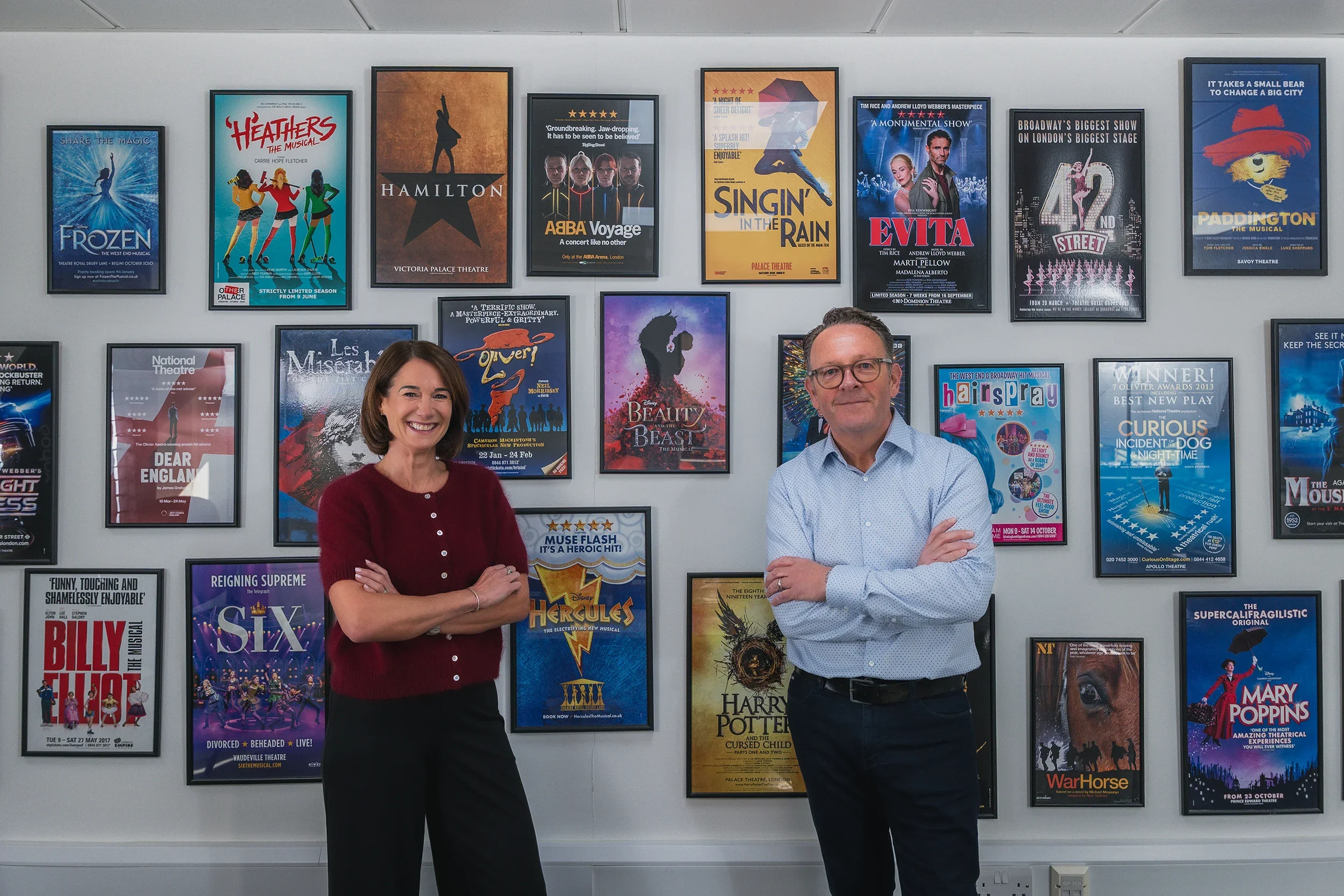A woman and a man standing in front of a wall decorated with framed theater posters, with the woman smiling and the man crossing her arms.